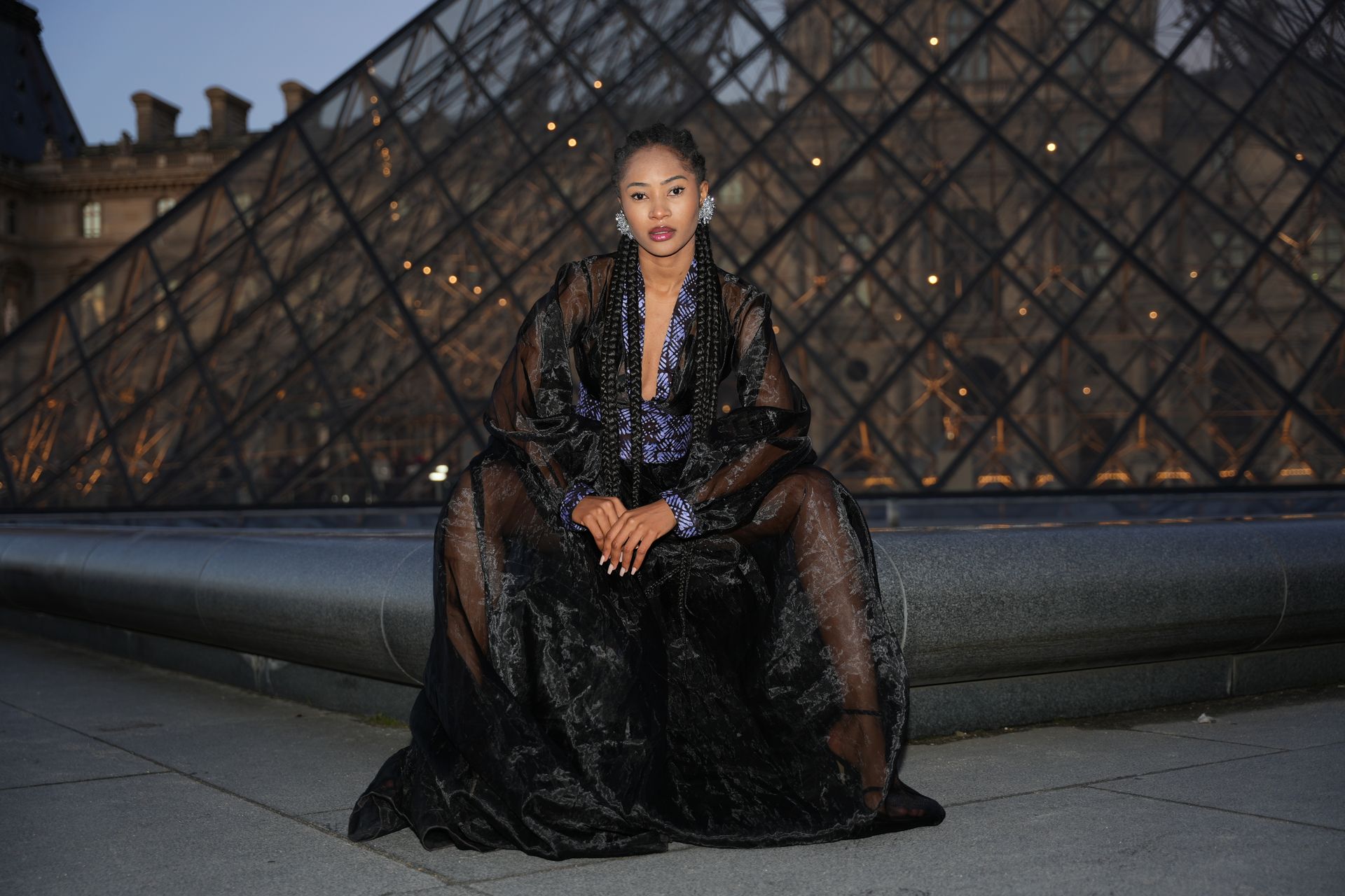 Woman in elegant dress poses by the illuminated Louvre Pyramid at twilight in Paris.