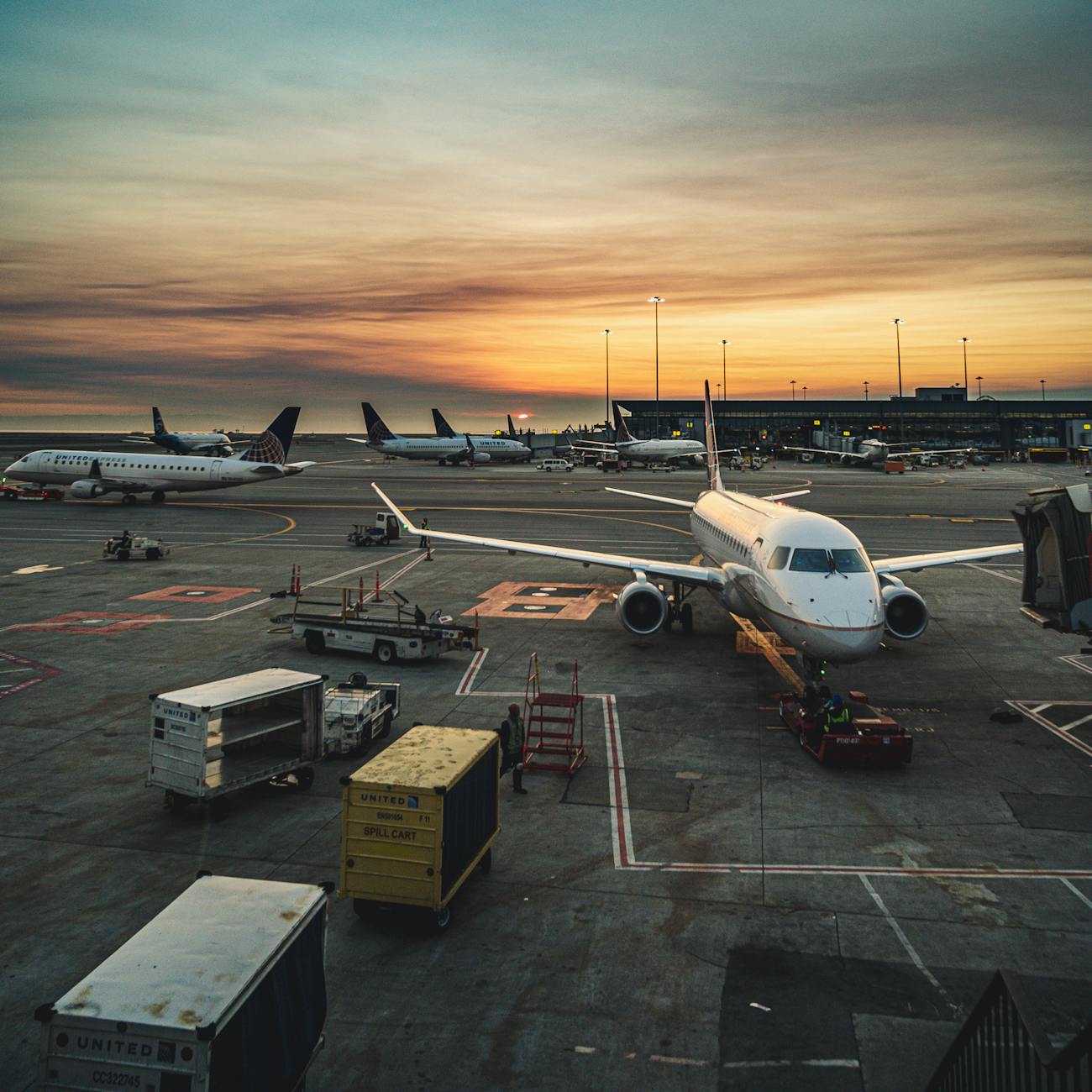 View of an airport tarmac at sunset with aircraft parked and cargo handling equipment in operation.