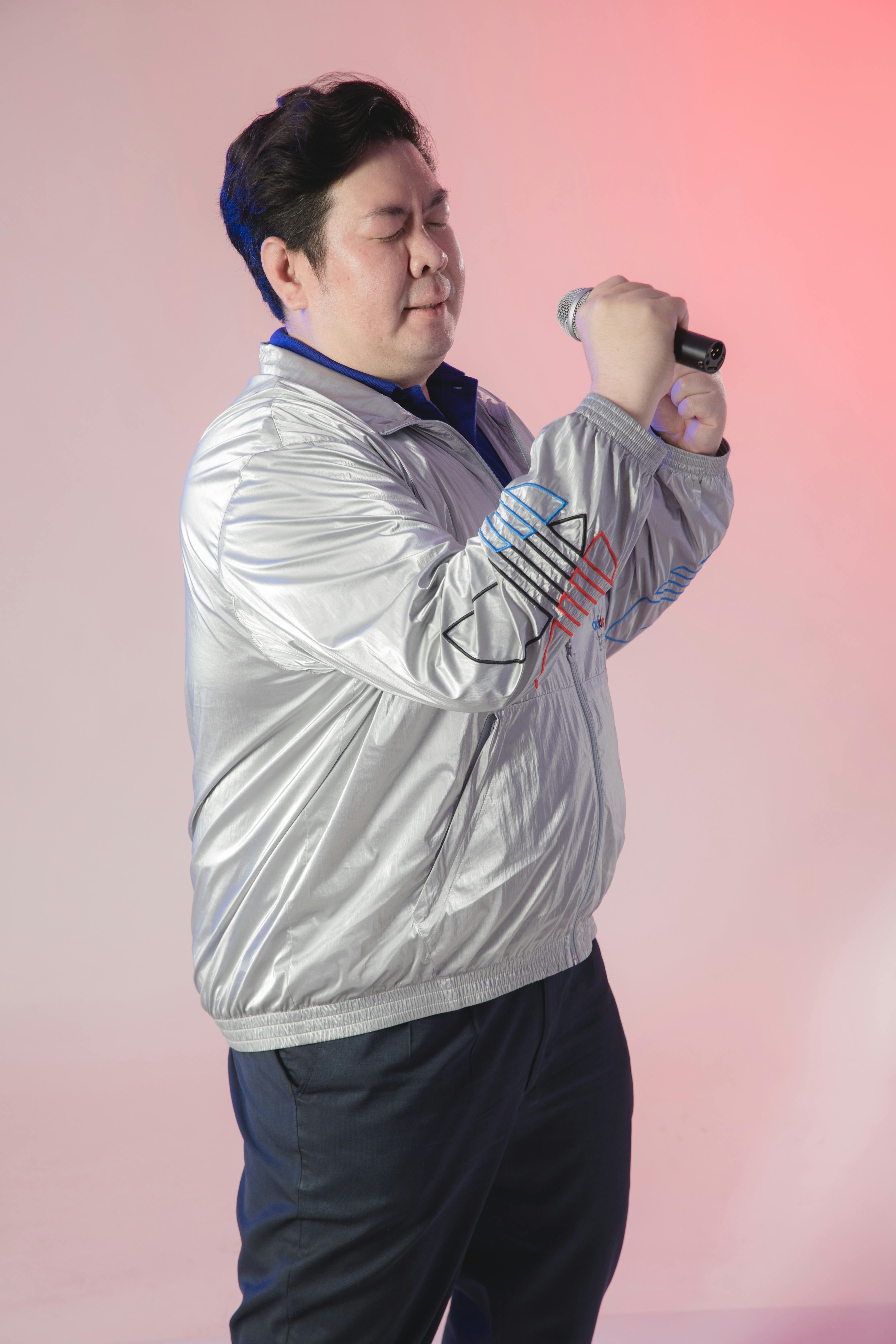 Asian man wearing a silver jacket passionately singing into a microphone.