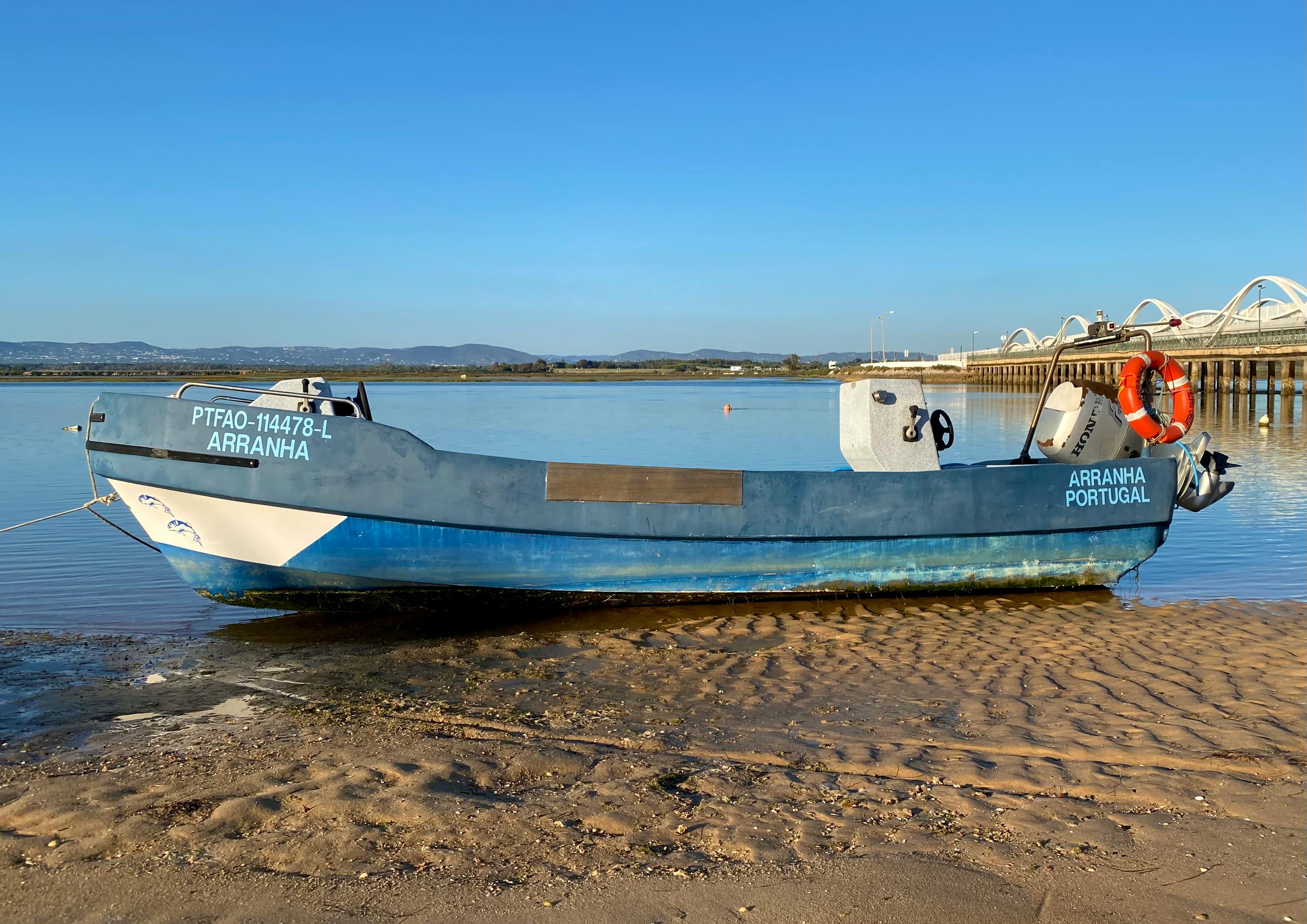 A blue fishing boat named Arranha resting on the sandy shore in Faro, Portugal.