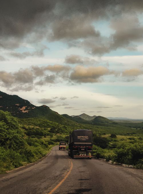 Free Two trucks drive along a scenic mountain road under a dramatic sky at dusk, surrounded by lush greenery. Stock Photo