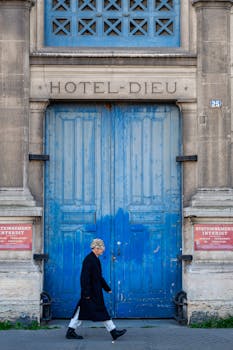 An elderly woman walks by the iconic blue doors of Hotel Dieu, set in a classical architectural facade.