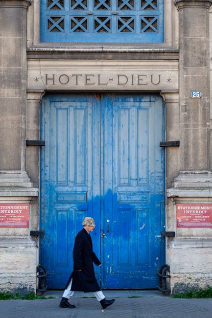 An elderly woman walks by the iconic blue doors of Hotel Dieu, set in a classical architectural facade.