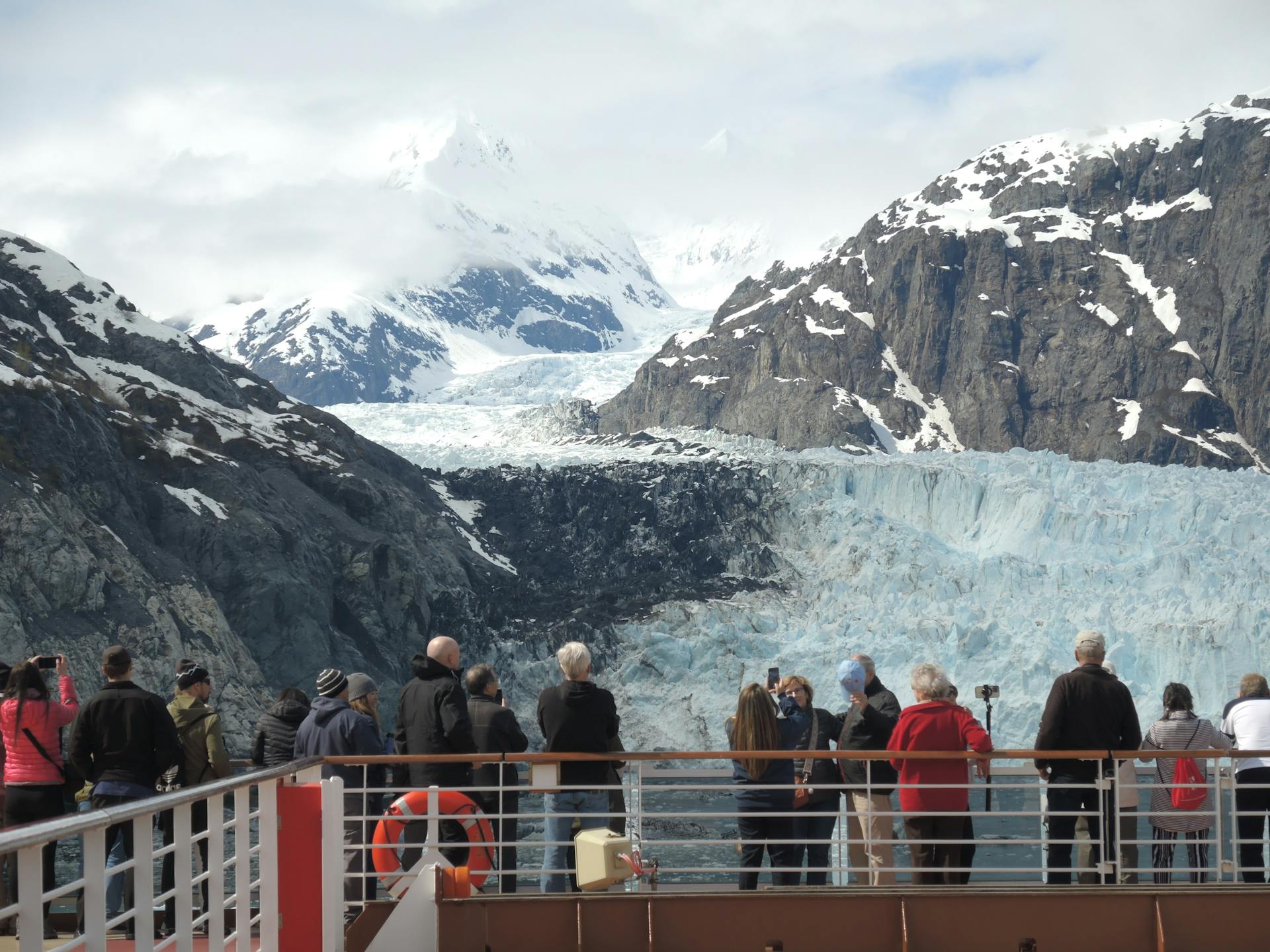 Tourists admire a stunning glacier from a cruise ship deck, surrounded by snowy mountains.