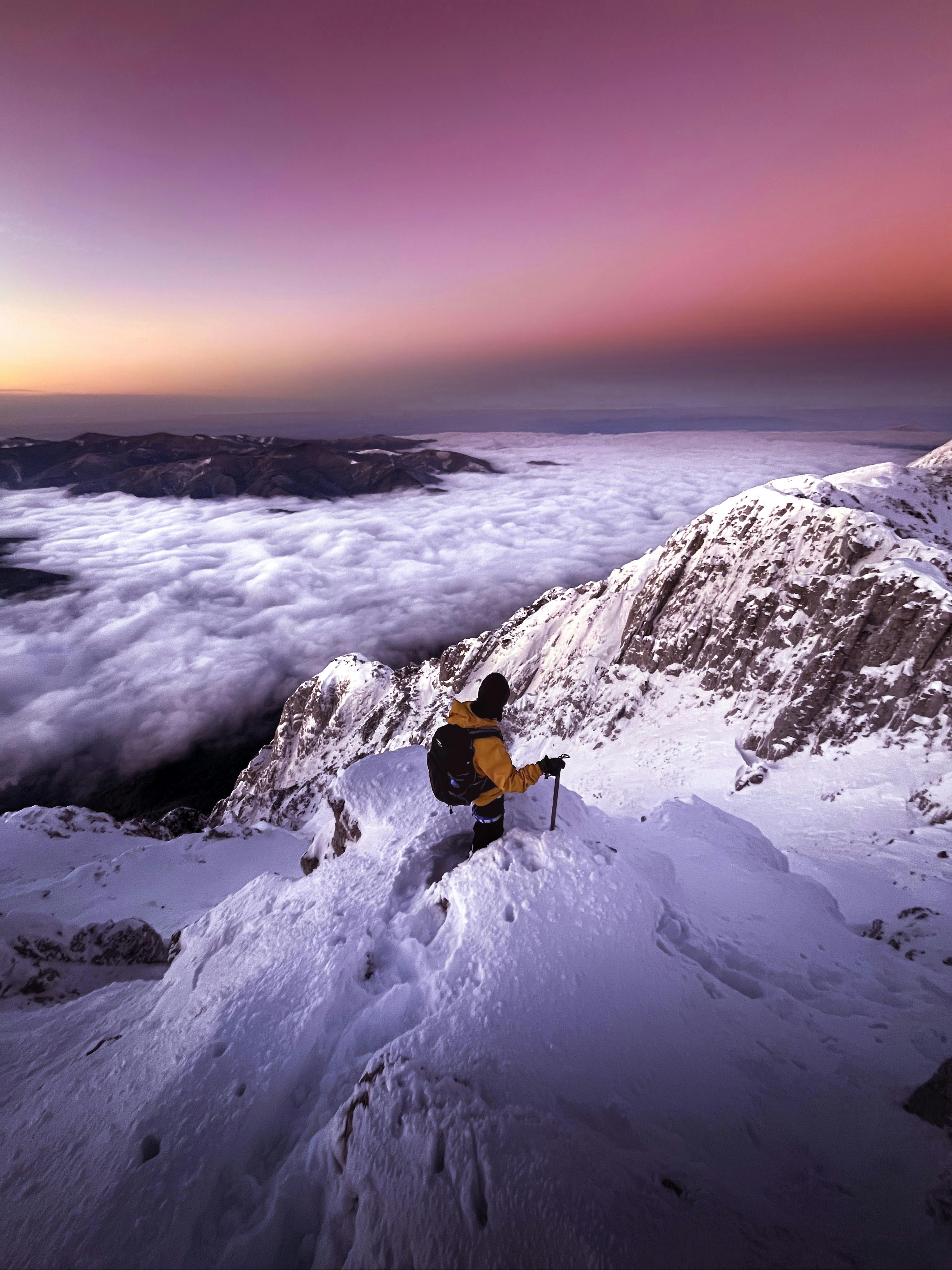 A lone mountaineer stands atop a snowy peak, enjoying a mystical sunset with snow-covered mountains and a violet sky.
