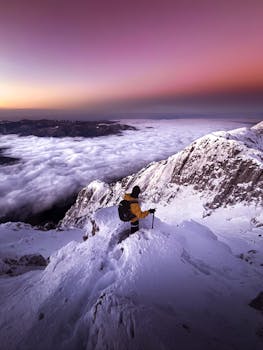 A lone mountaineer stands atop a snowy peak, enjoying a mystical sunset with snow-covered mountains and a violet sky.