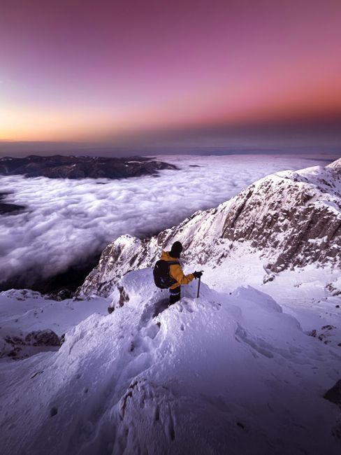 A lone mountaineer stands atop a snowy peak, enjoying a mystical sunset with snow-covered mountains and a violet sky.