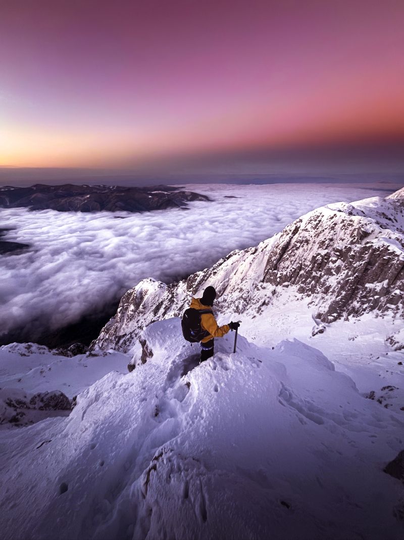 A lone mountaineer on a snowy peak glowing in sunset alpenglow