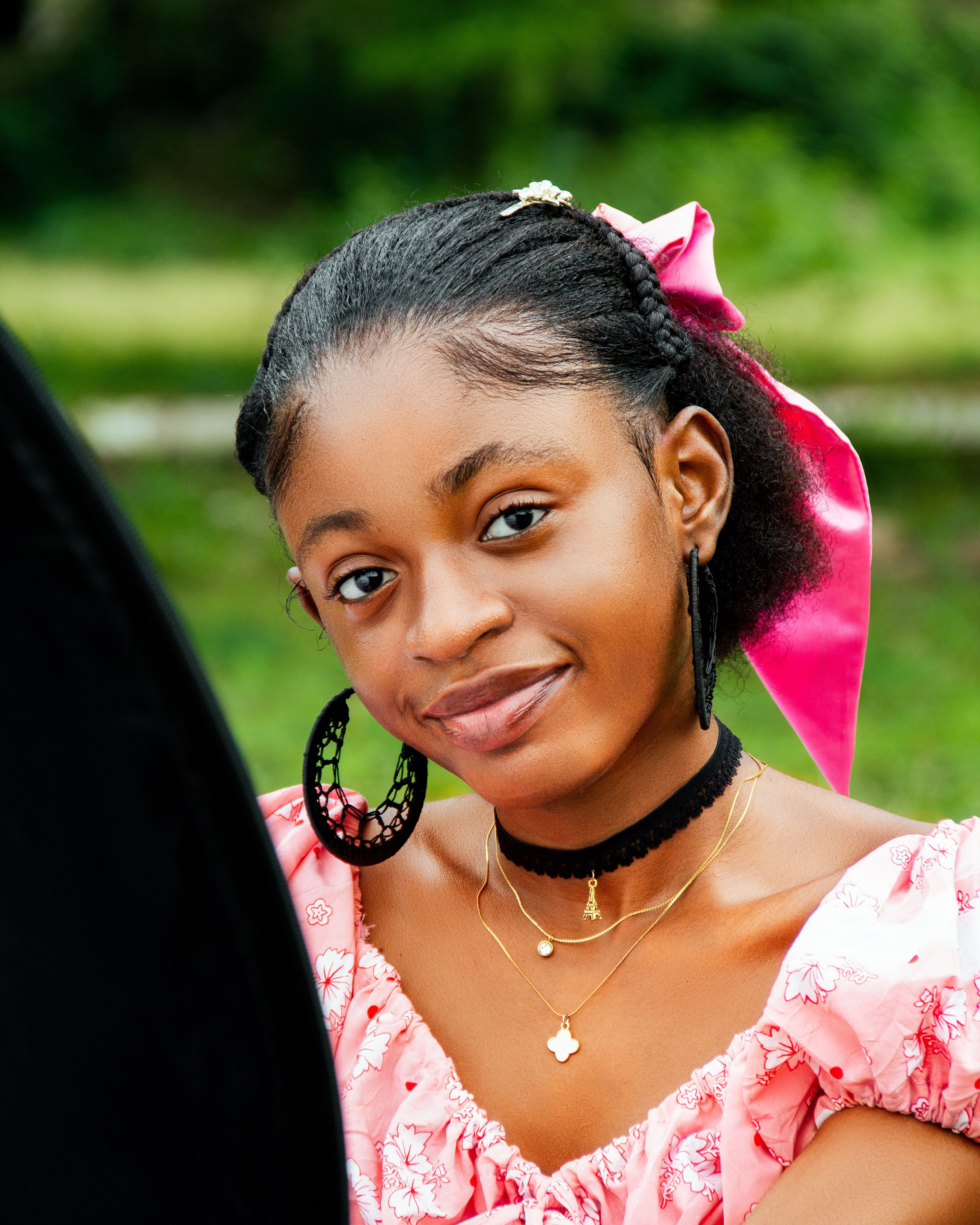 Smiling woman in pink dress with bow in hair and hoop earrings outdoors.