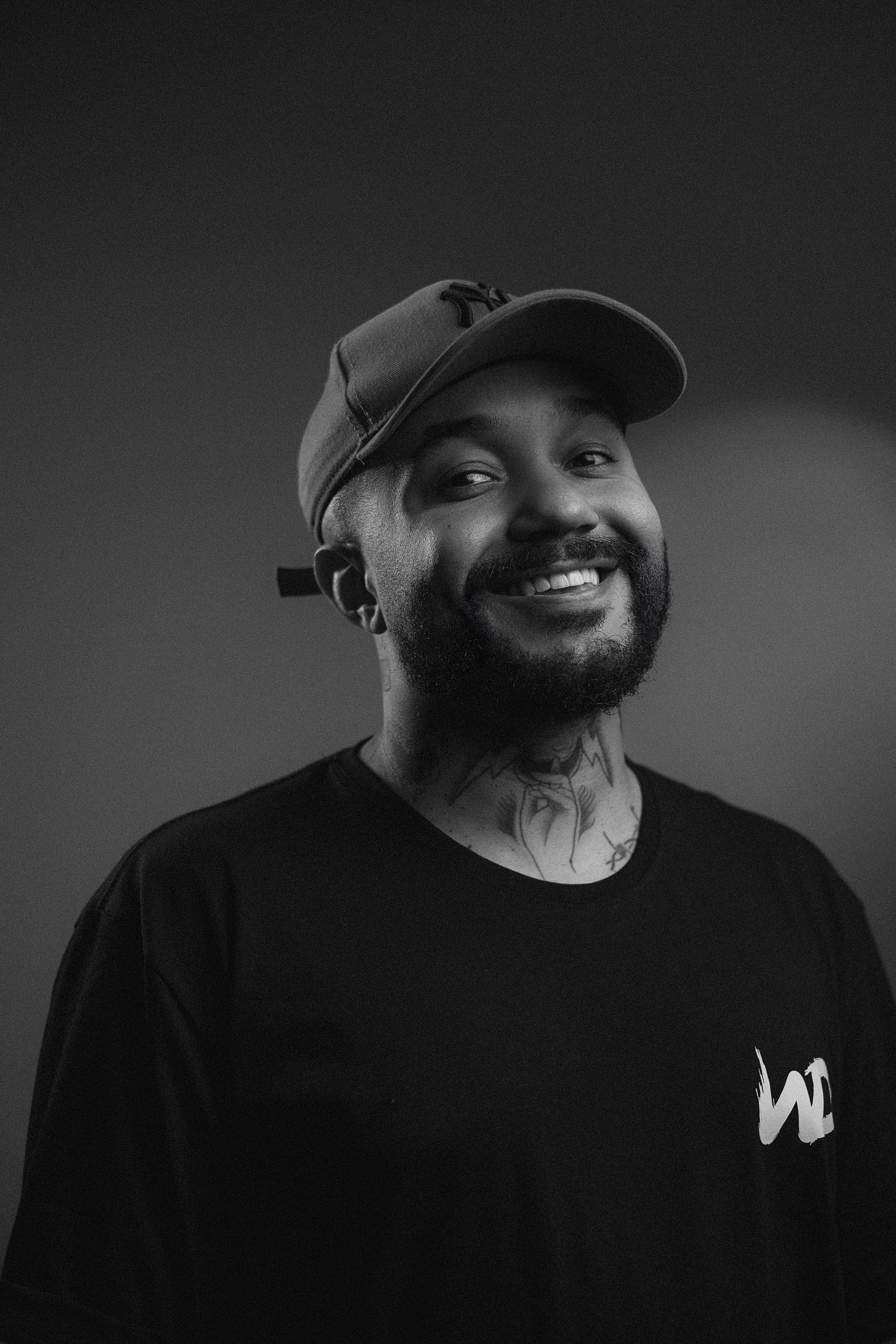 A black and white portrait of a smiling man wearing a cap and T-shirt.