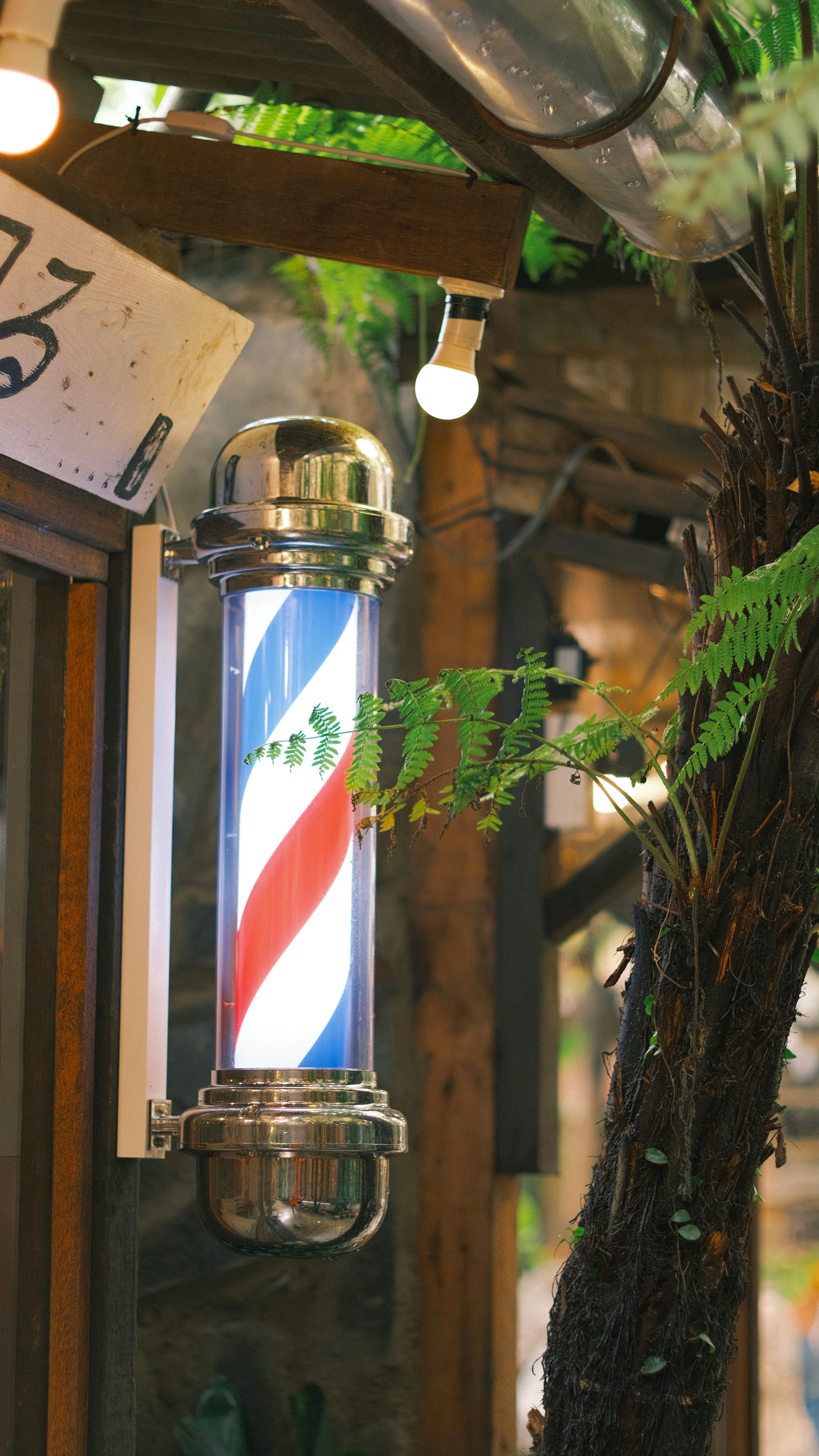 Classic barber pole outside shop with greenery in view. Bright and inviting.