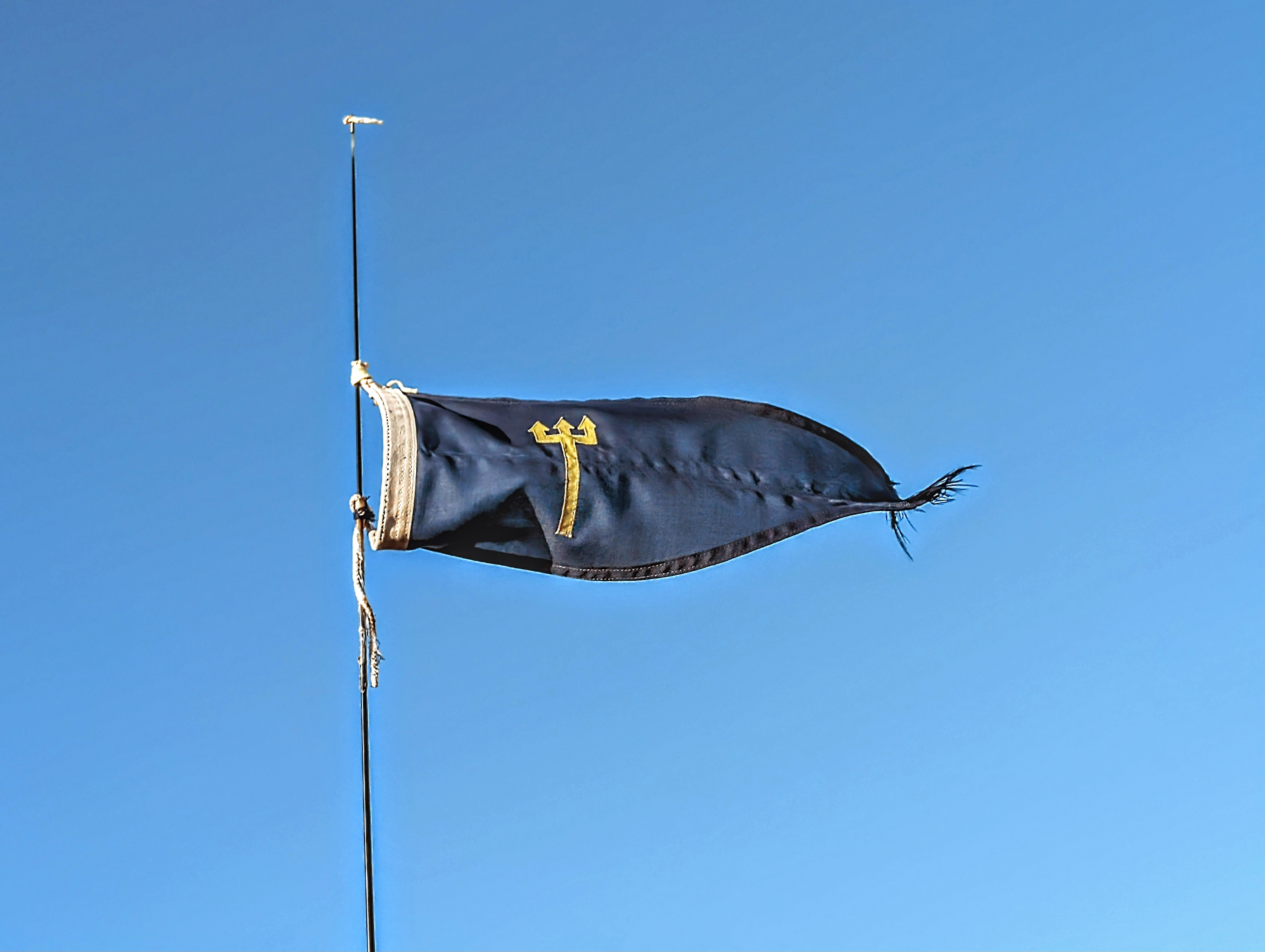 A navy blue flag with a yellow symbol flutters against a clear blue sky, symbolizing identity or event.