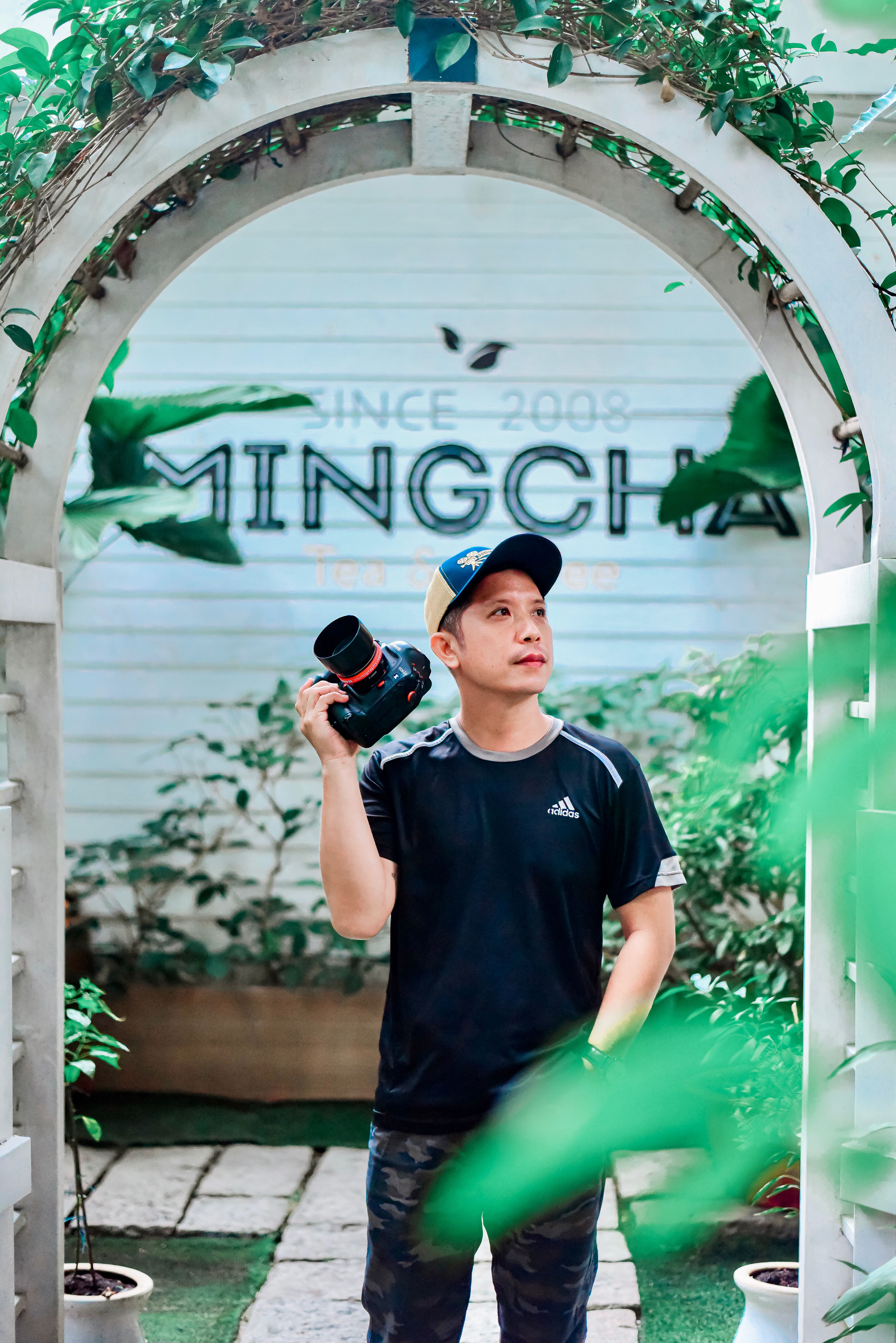 A young male photographer stands under an arch holding a camera amidst greenery.