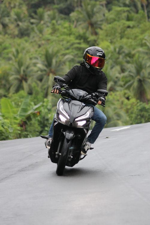 Free A motorcyclist in protective gear rides a scooter on a tropical road, surrounded by lush greenery. Stock Photo