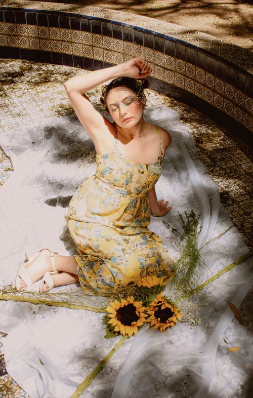 Free A woman in a floral yellow dress poses with sunflowers in a tiled courtyard, capturing an artistic and vibrant summer vibe. Stock Photo
