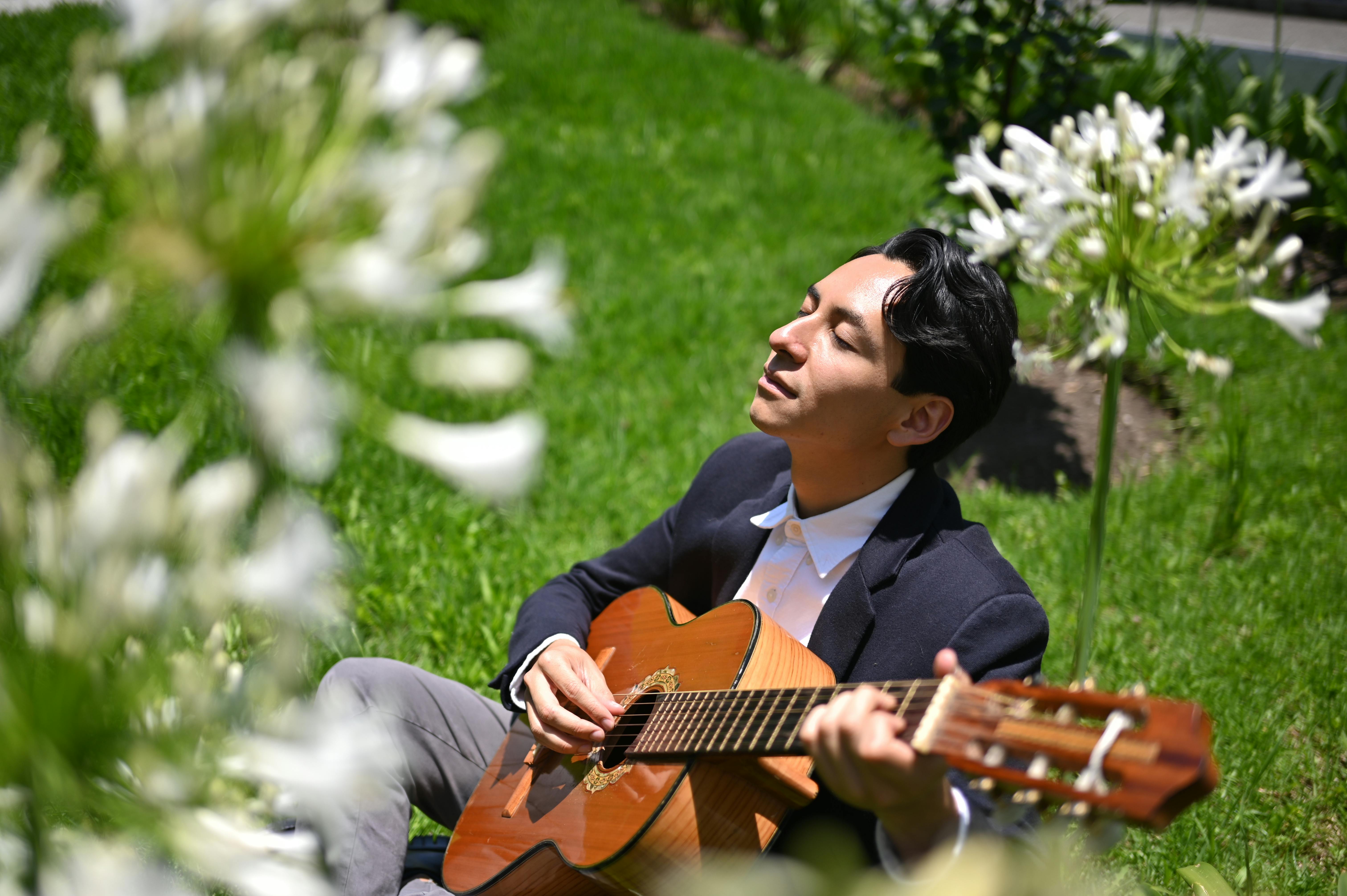 Young man enjoys playing guitar in a sunny garden, surrounded by white flowers and green grass.
