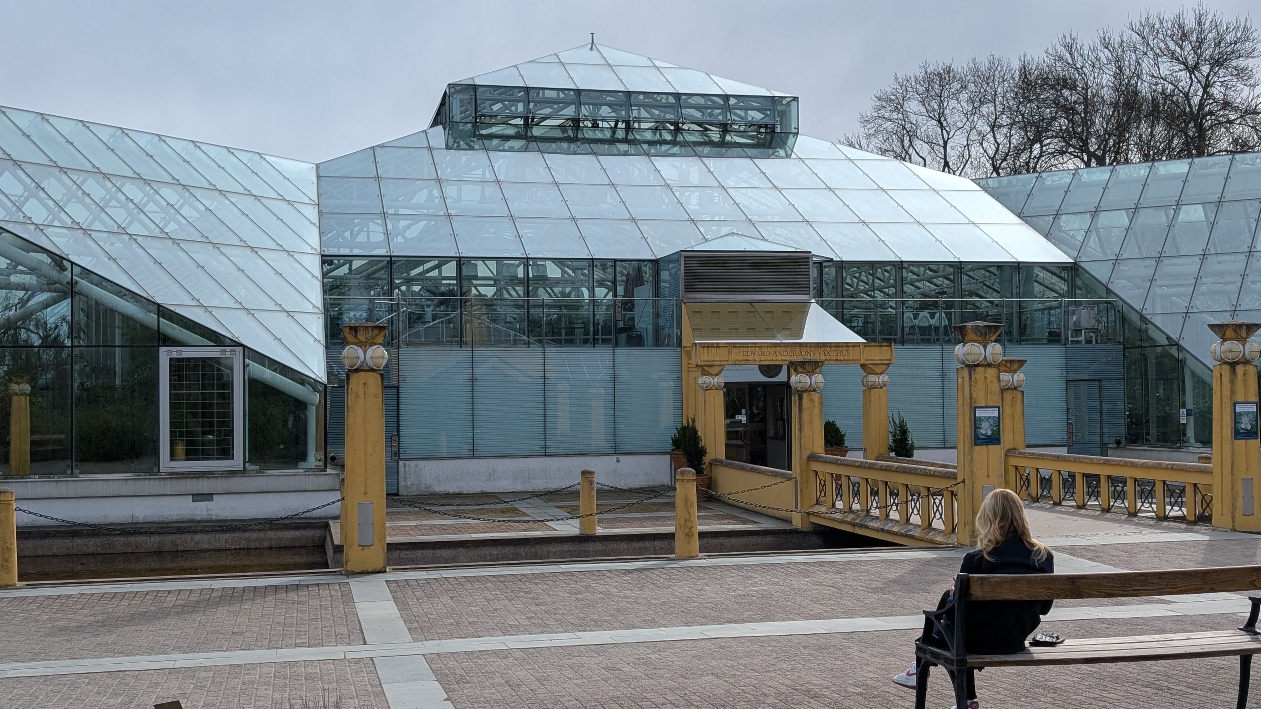 A woman sits on a bench near a glass building with yellow columns, perfect for urban architecture themes.