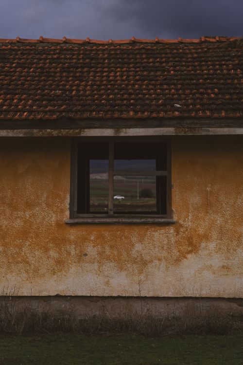 Free A weathered rural house with a window reflecting a scenic landscape, under a moody sky. Stock Photo
