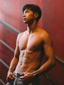 Athletic shirtless man posing confidently by a red wall, showcasing toned muscles and fitness.