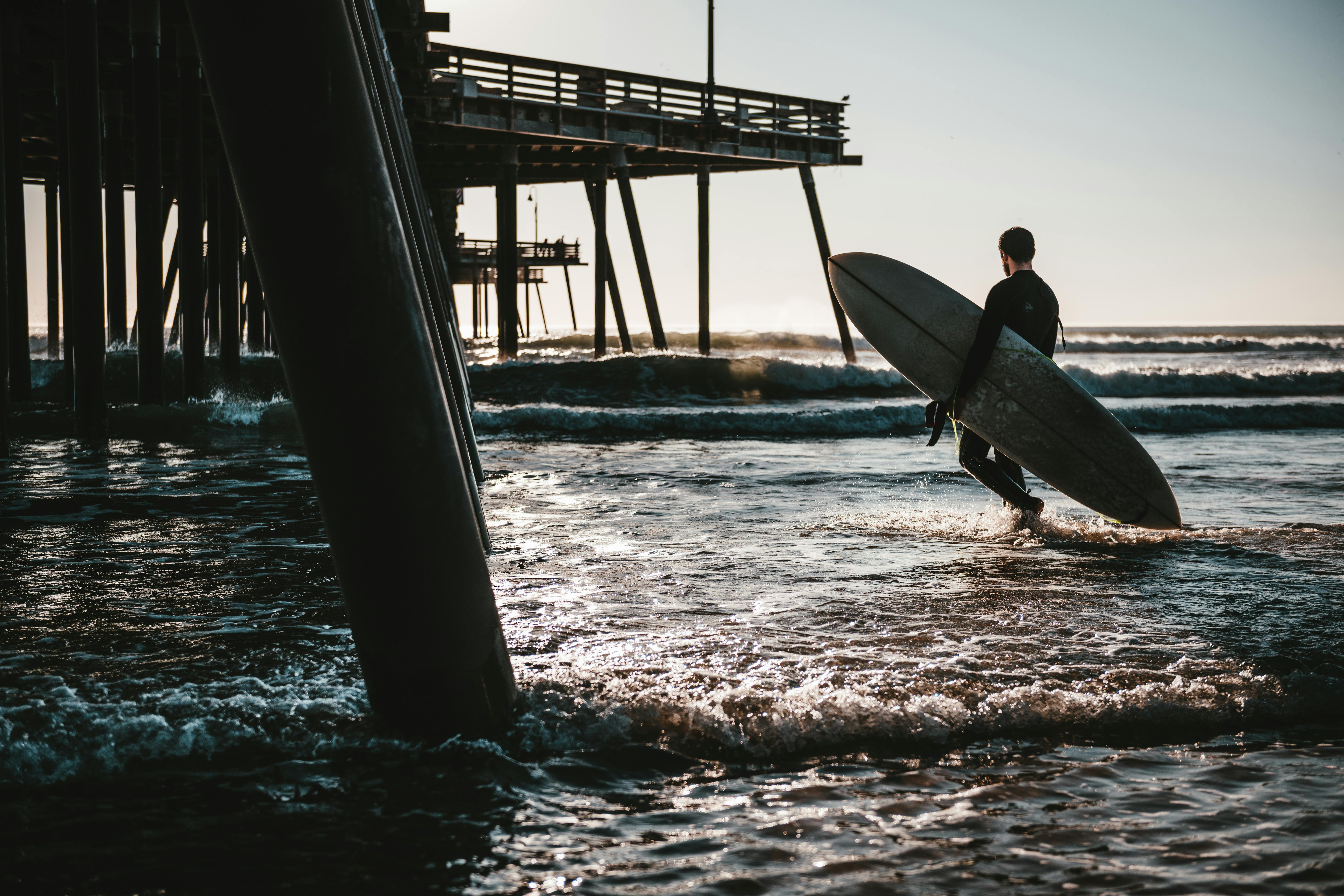 A surfer walks with a surfboard by the pier at Pismo Beach, California, highlighting a serene sunset scene.