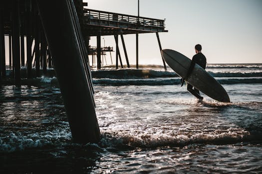 A surfer walks with a surfboard by the pier at Pismo Beach, California, highlighting a serene sunset scene.