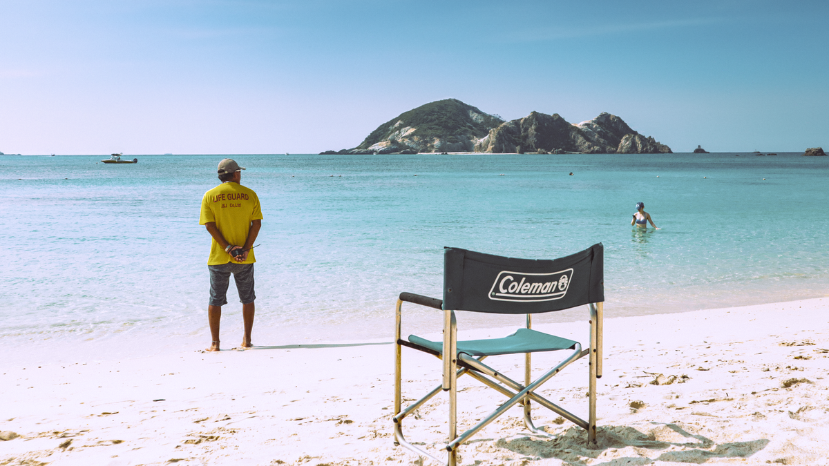 A white sand beach on Tokashiki with turquoise shallows, an empty green camp chair in the foreground, a lifeguard in a yellow LIFE GUARD shirt watching the water, a small rocky island offshore, and one swimmer.