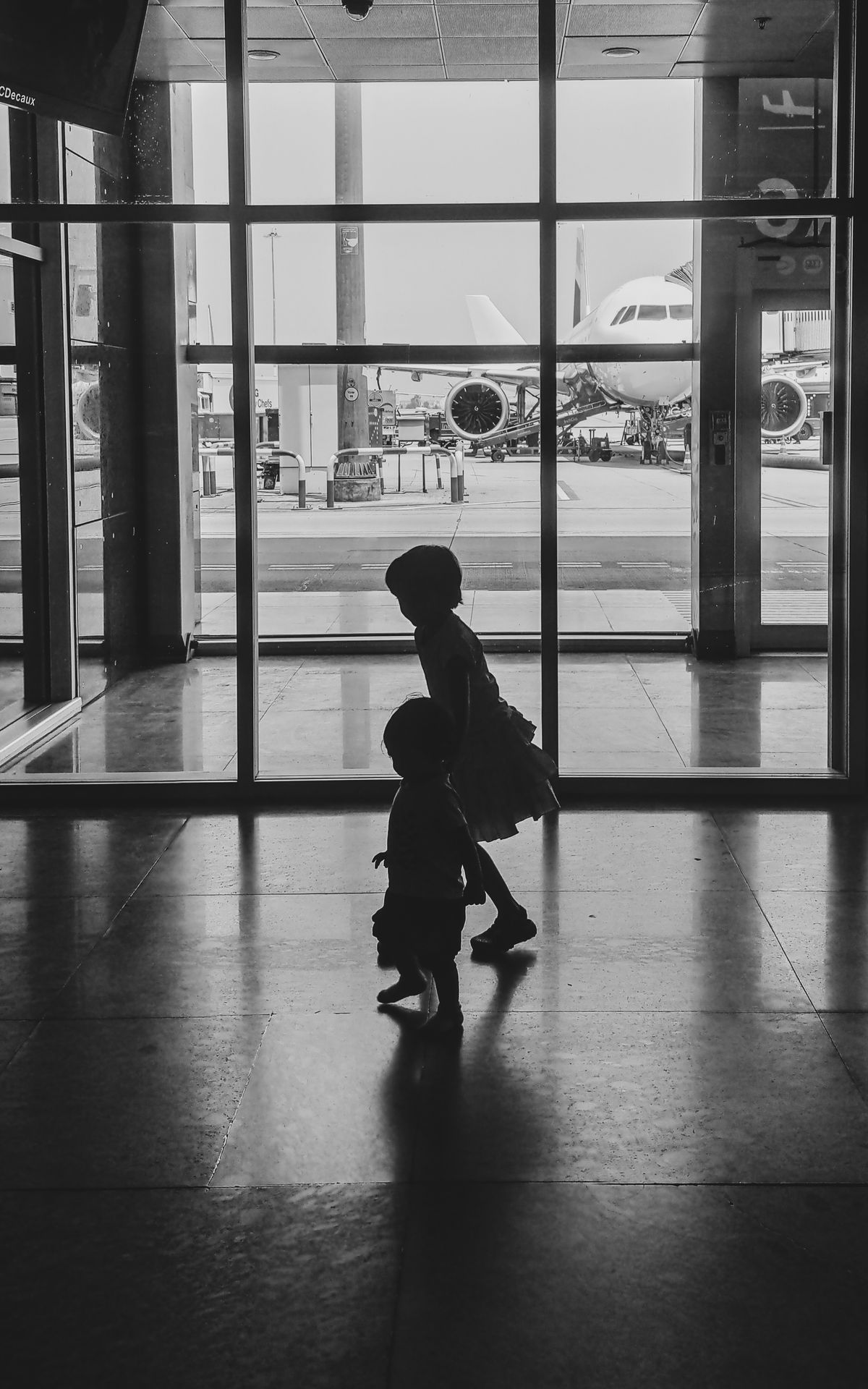 Silhouettes of two young children walking through a terminal towards the gate, with an aircraft visible outside