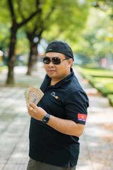 Man in sunglasses and cap holds currency notes, standing outdoors on a tree-lined path.