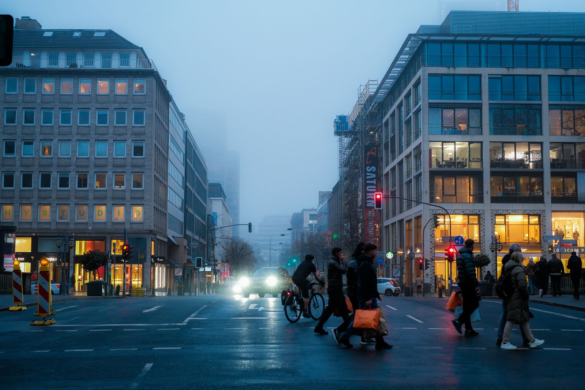 City intersection with people and cyclists in foggy evening, vibrant lights contrast the mist.