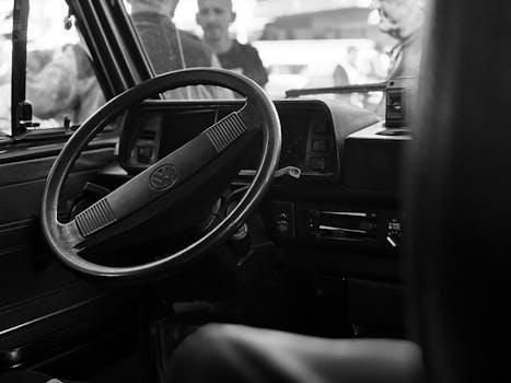 Black and white image of a vintage car interior featuring a steering wheel with blurred people outside.