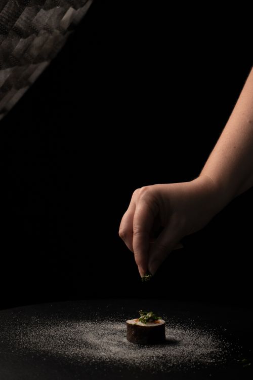 Free A hand delicately sprinkles garnish on sushi in low light setting, creating an artistic culinary scene. Stock Photo