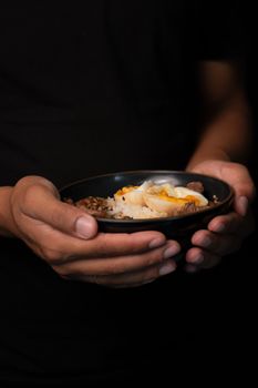 Close-up of hands holding a rice bowl with a perfectly poached egg on top, set against a dark background.