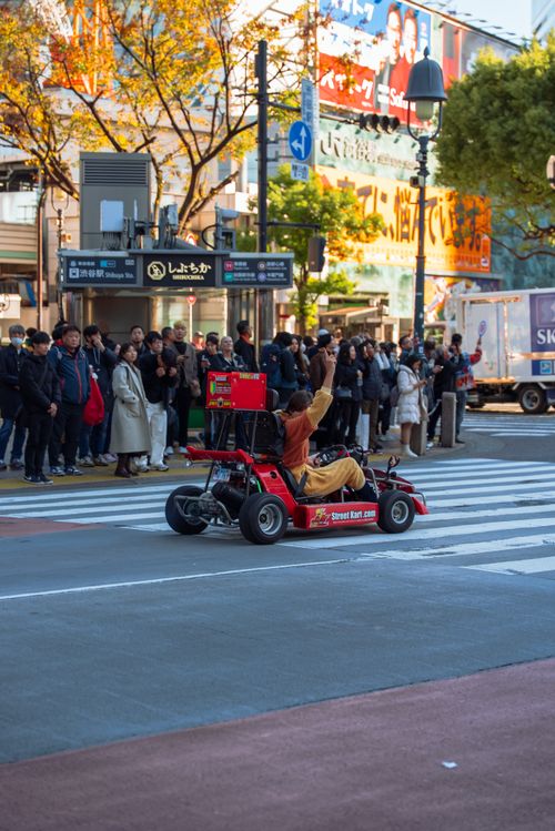 Free Go-kart driver in urban intersection with crowd watching on sidewalk, vibrant city scene. Stock Photo