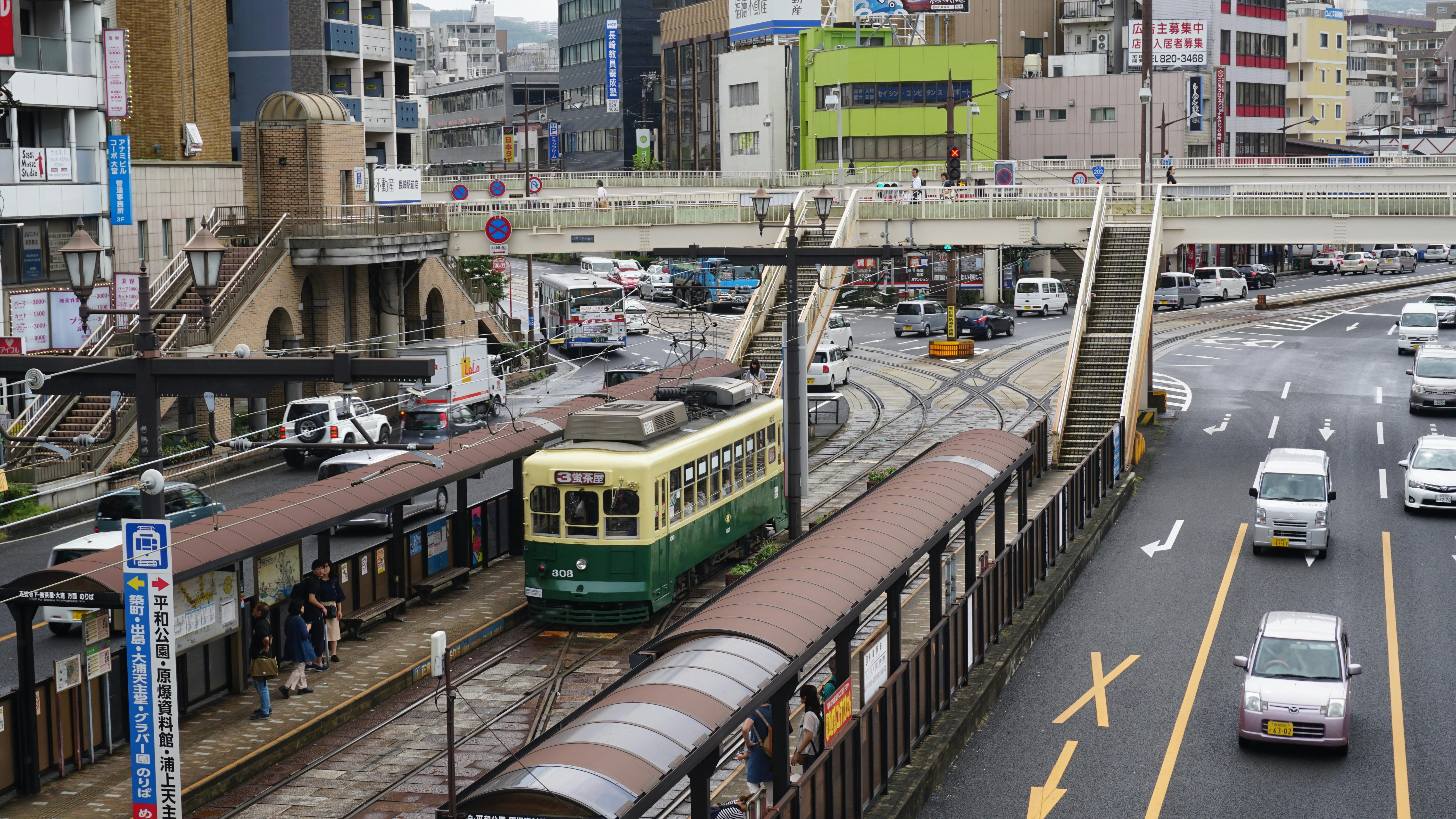 Urban scene in Nagasaki City featuring a tram and bustling street traffic with a pedestrian overpass.