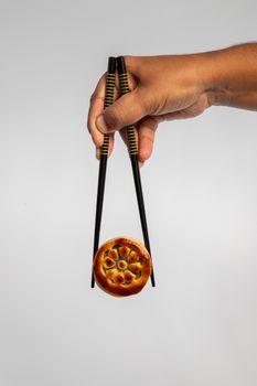 A hand holding a traditional mooncake using chopsticks on a simple background.