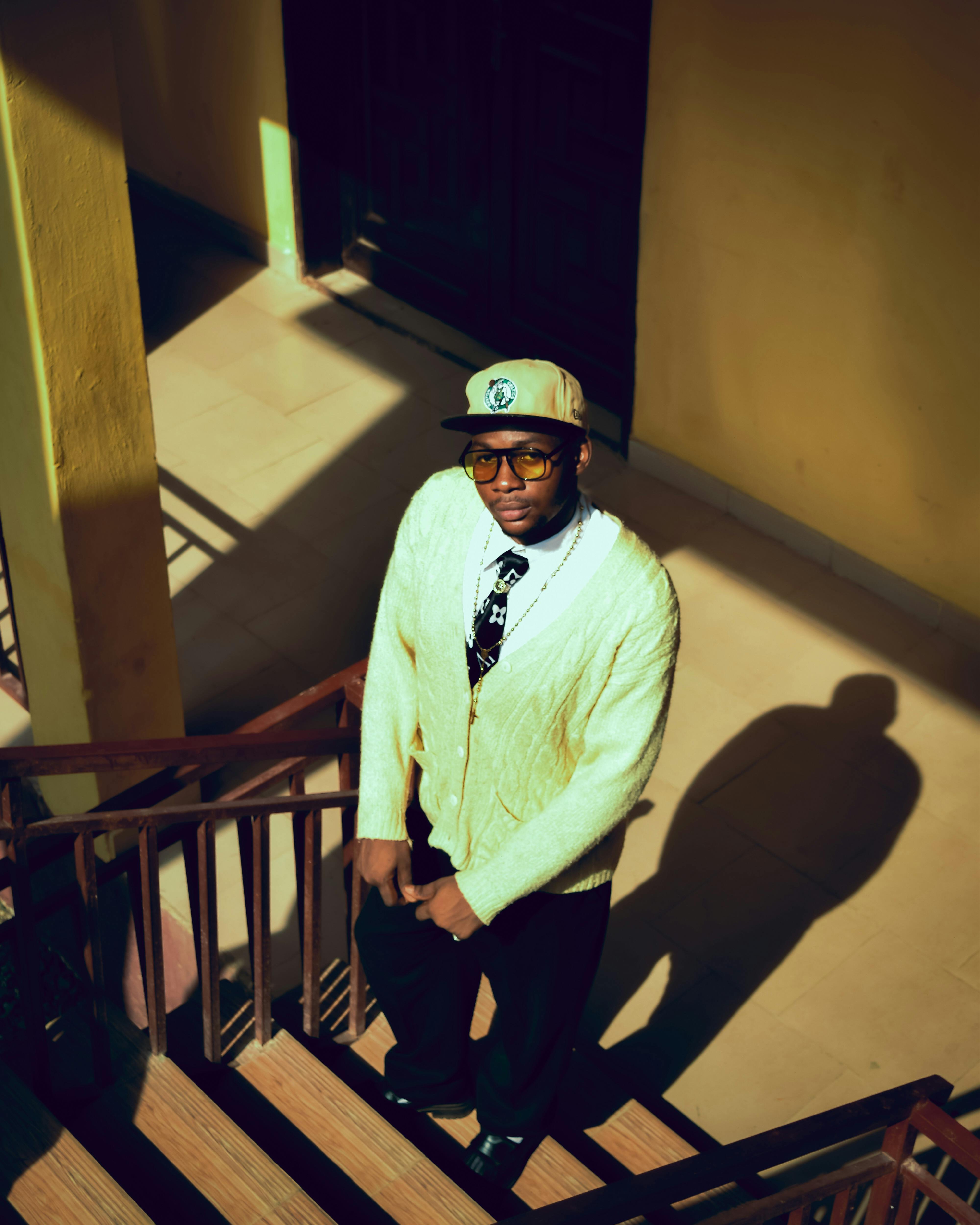 A fashionable man stands on an outdoor staircase, casting a shadow in bright sunlight.