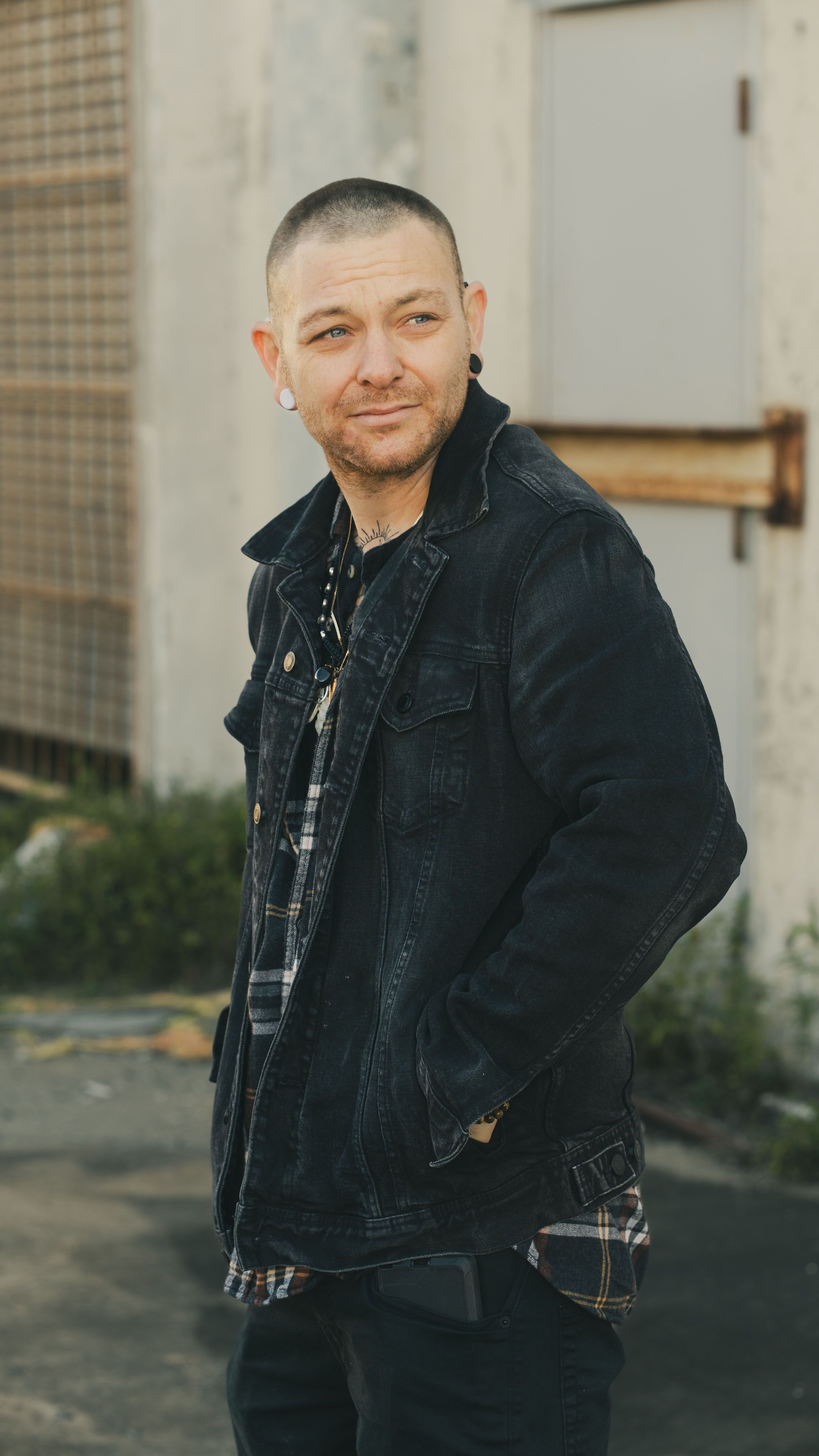 A stylish man in a denim jacket poses outdoors against an industrial backdrop, exuding casual confidence in Houston, Texas.