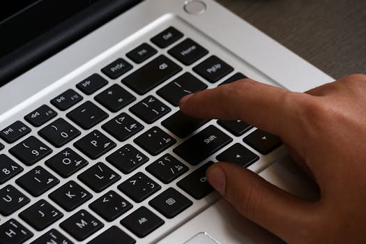 Close-up image of a hand typing on a laptop keyboard featuring Arabic script. Modern workspace setup.