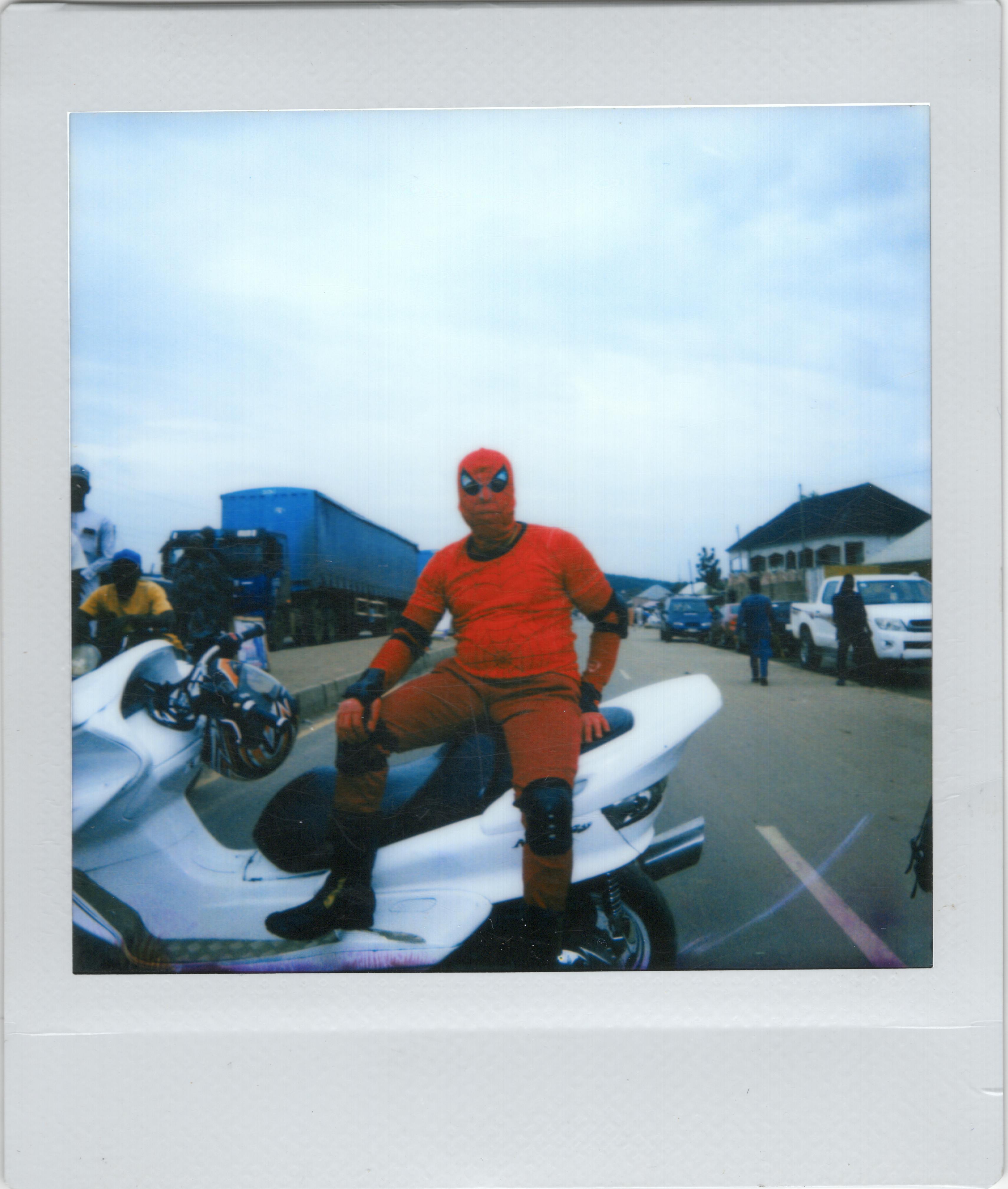 Man dressed as superhero sitting on a white motorcycle in an open road setting.