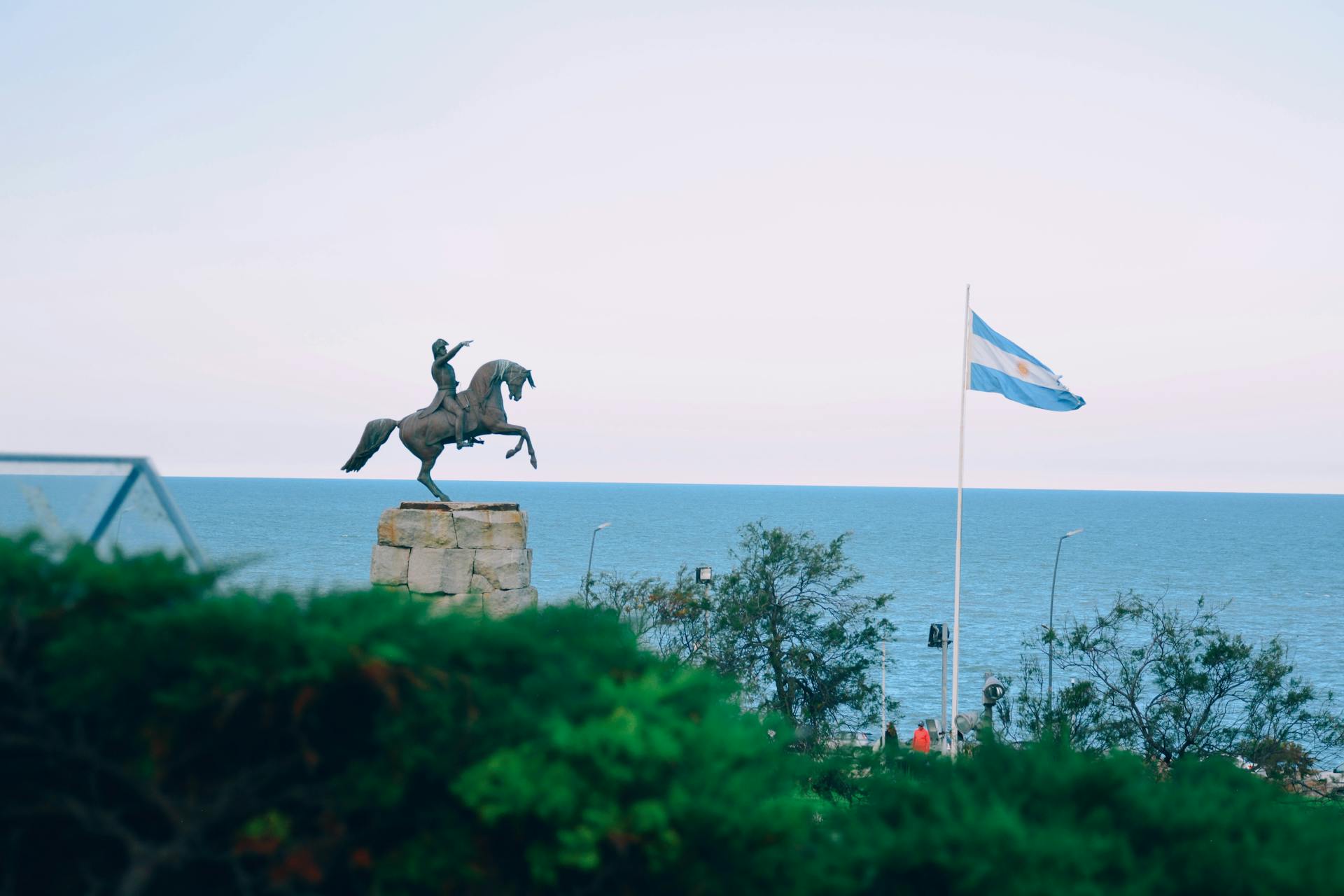 Equestrian statue with Argentine flag by the sea on a clear day.