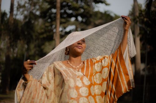 Free An African woman gracefully lifts a lace veil while wearing colorful traditional attire in a serene outdoor setting. Stock Photo
