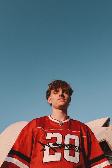 A young man wearing a sports jersey poses in front of the iconic Sydney Opera House under clear skies.