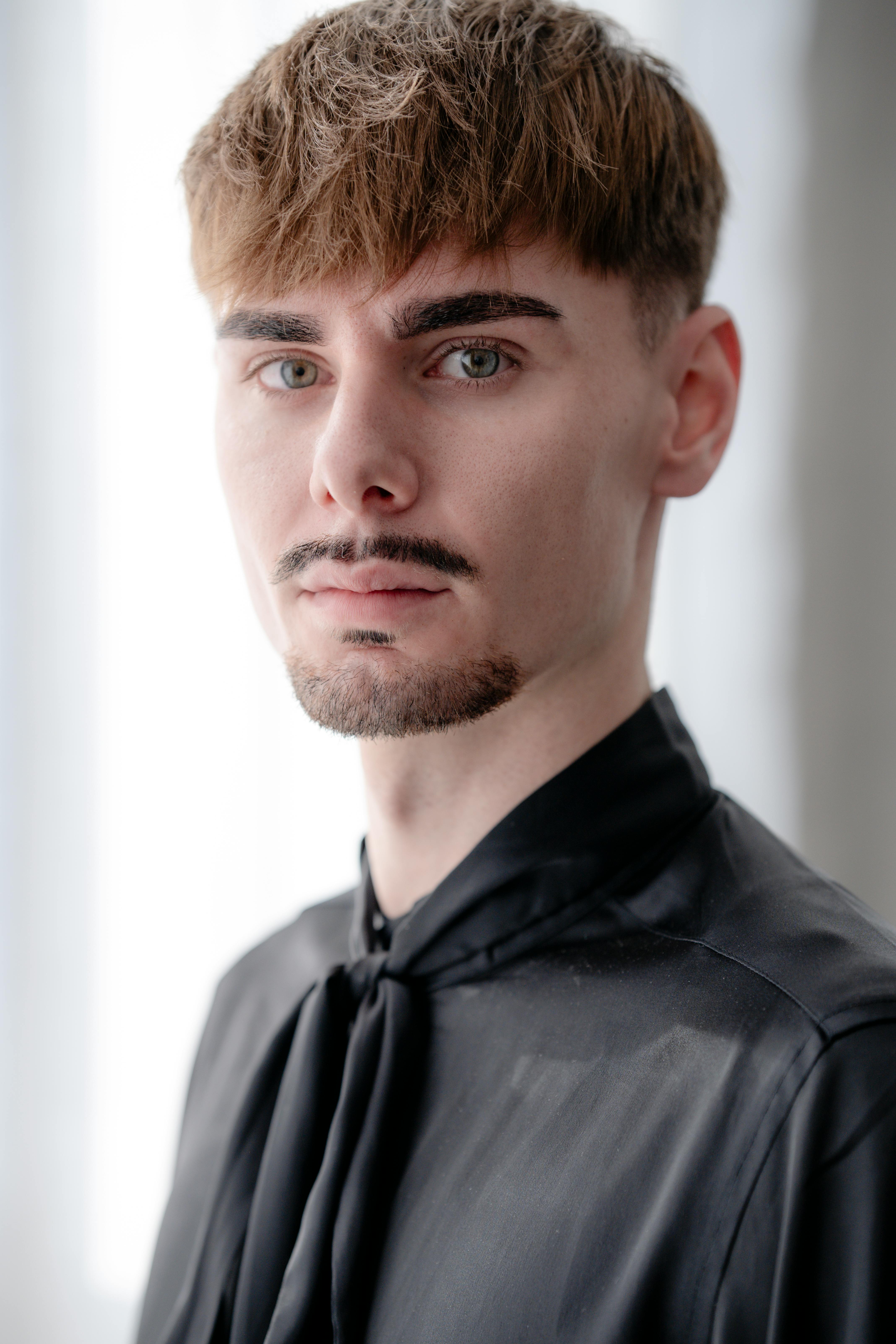 Close-up portrait of a young man with a mustache, wearing a black shirt indoors.