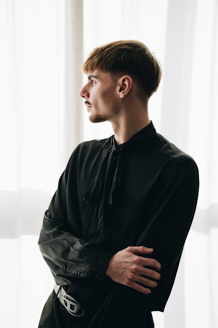 A young man in dark clothing gazes out a window in a contemplative mood.