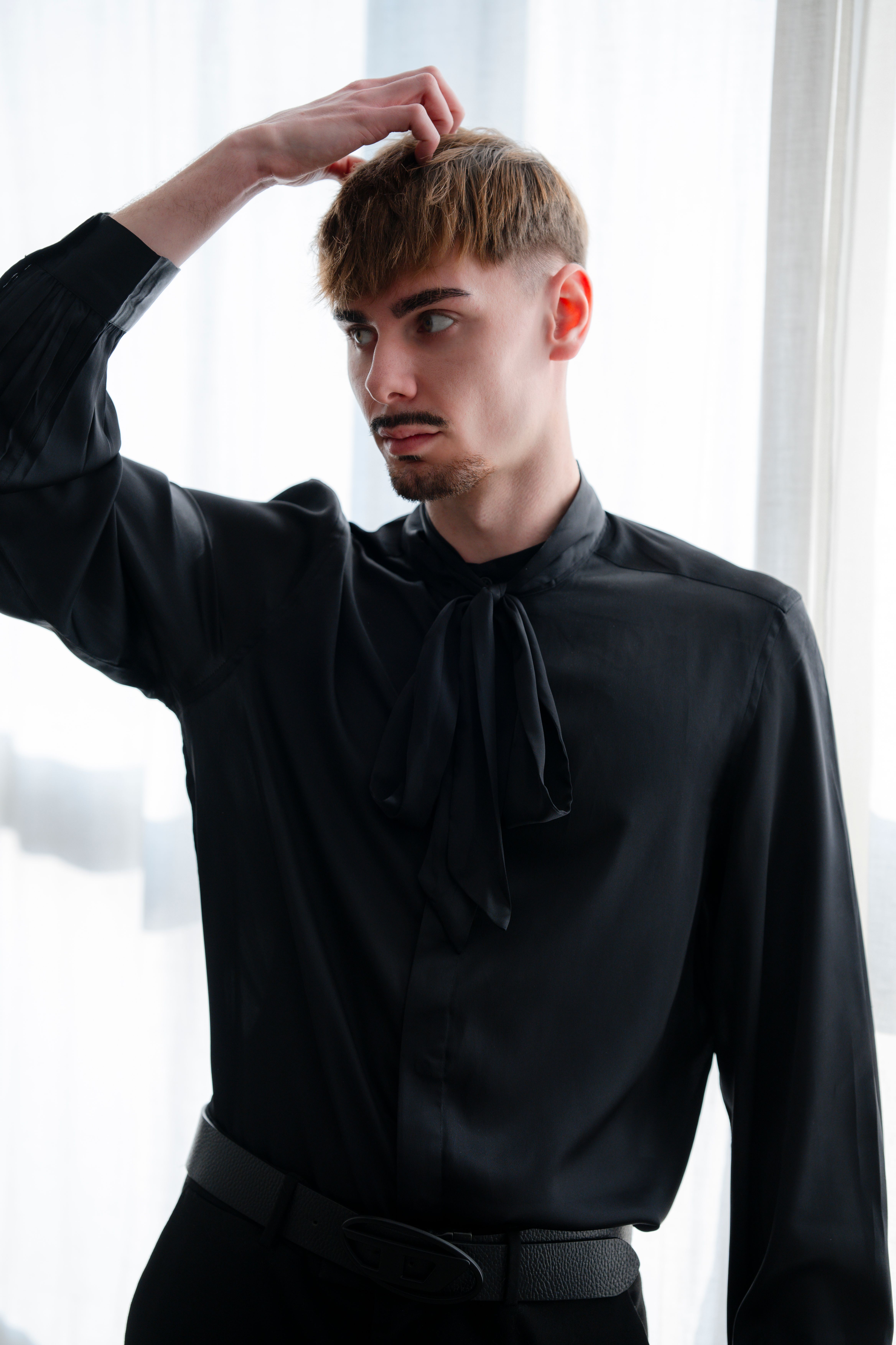 Stylish adult man in black shirt posing thoughtfully by window, natural light.