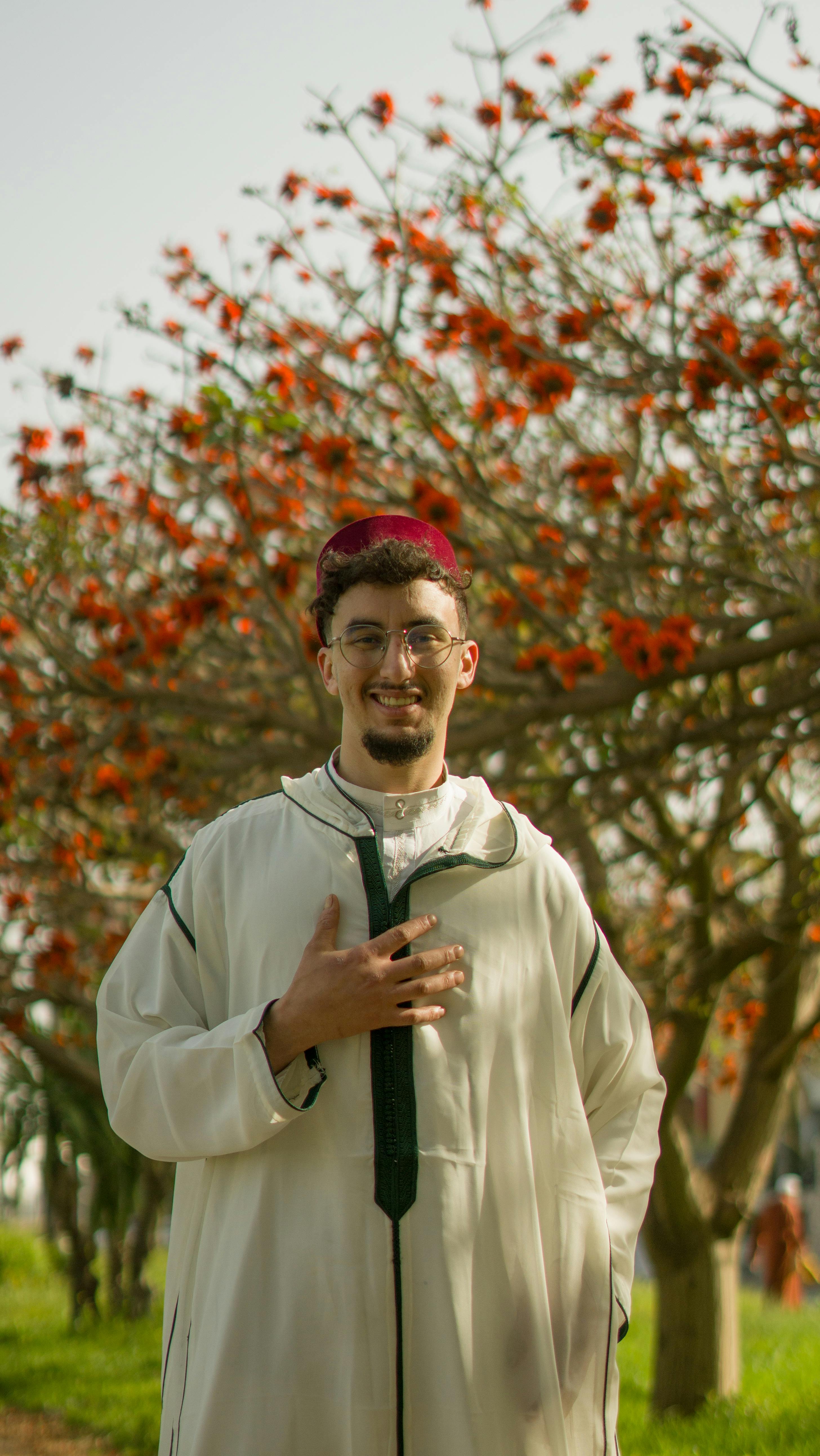 A man wearing traditional Moroccan attire stands amidst vibrant flowering trees on a sunny day.
