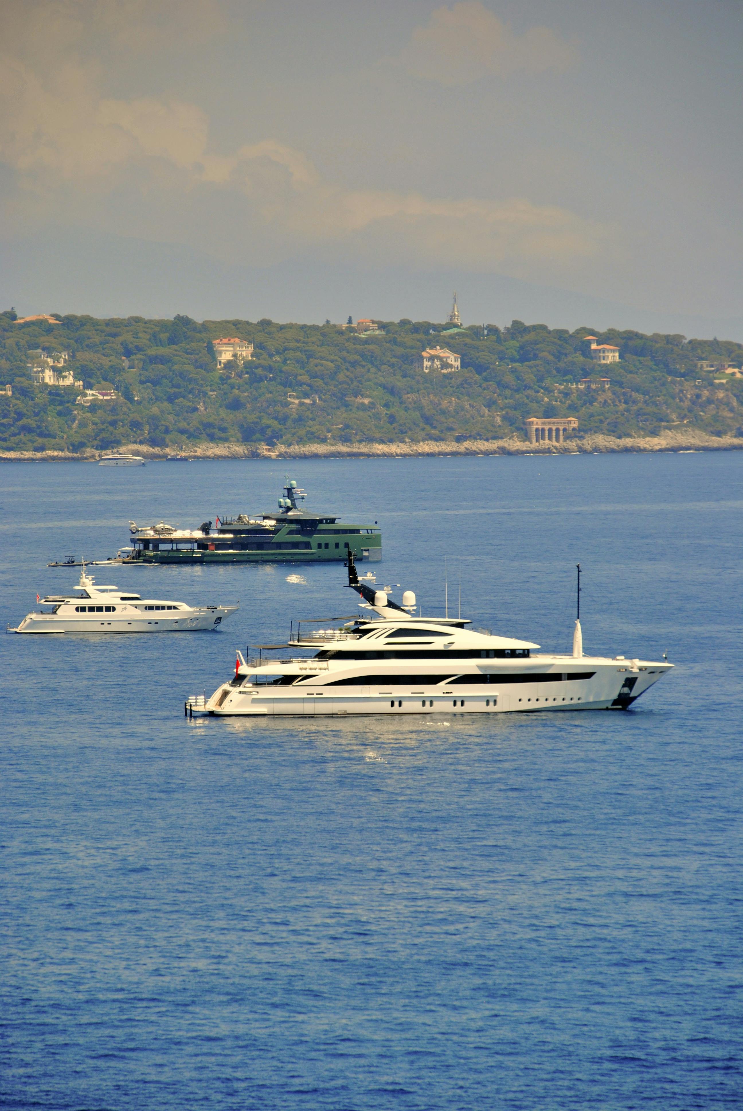 Three yachts on blue water, with greenery in the background under a cloudy sky.