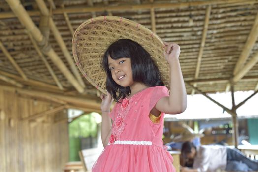 A young girl in a pink dress smiles while wearing a bamboo hat indoors.