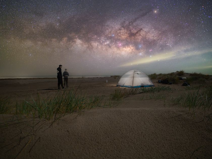 Two travellers stargazing beside a lit tent under the Milky Way