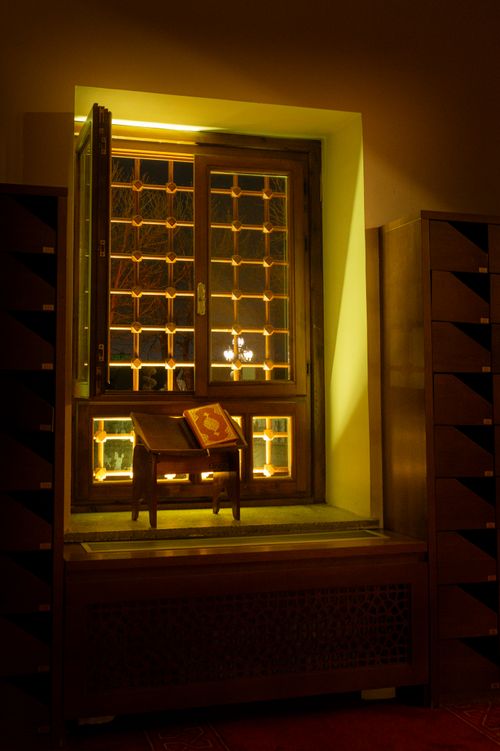 Free A beautifully lit mosque interior featuring an open Quran on a wooden stand by a window with intricate designs. Stock Photo
